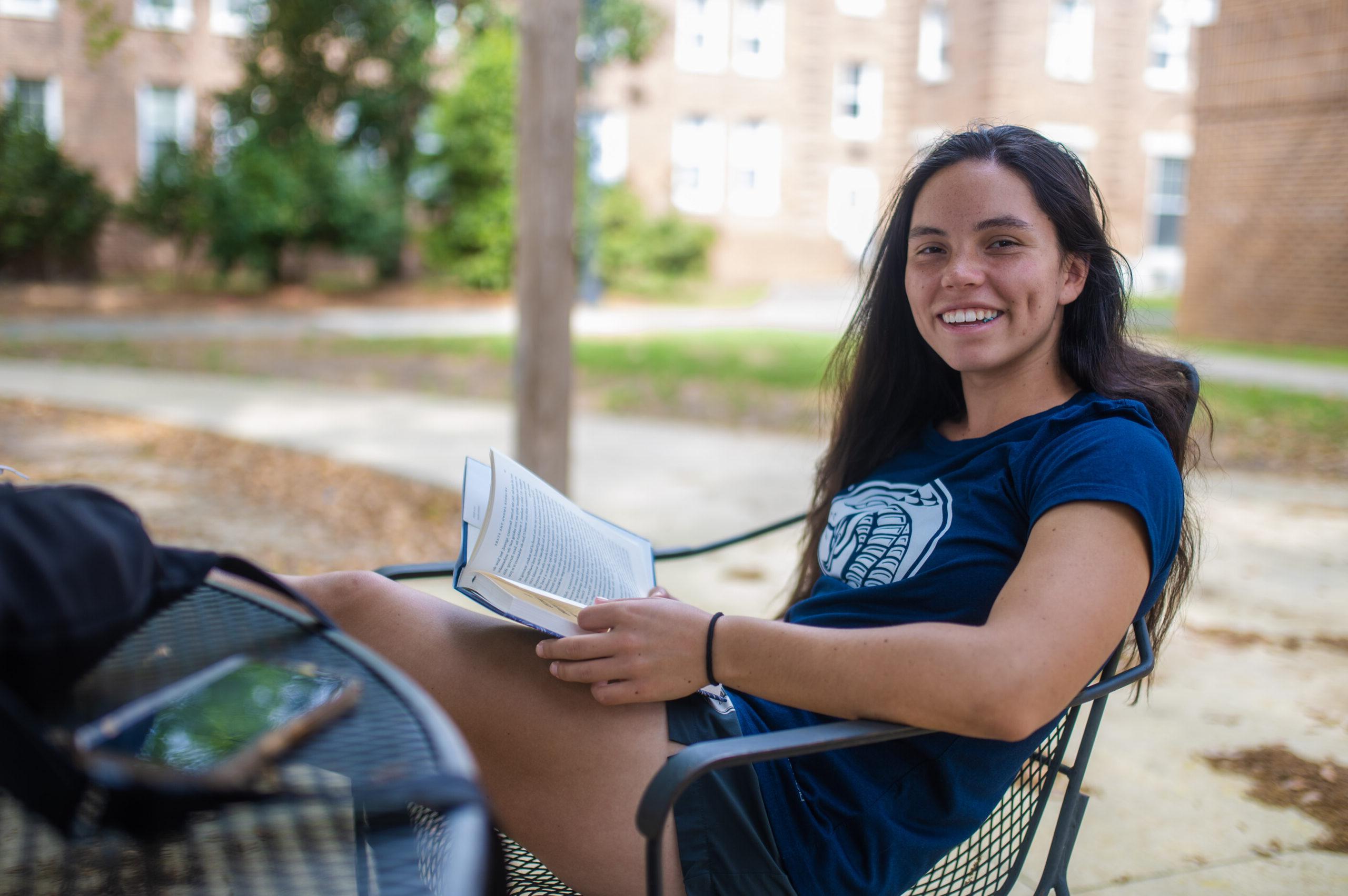 student lounges in a chair with a book in her hand smiling at the camera